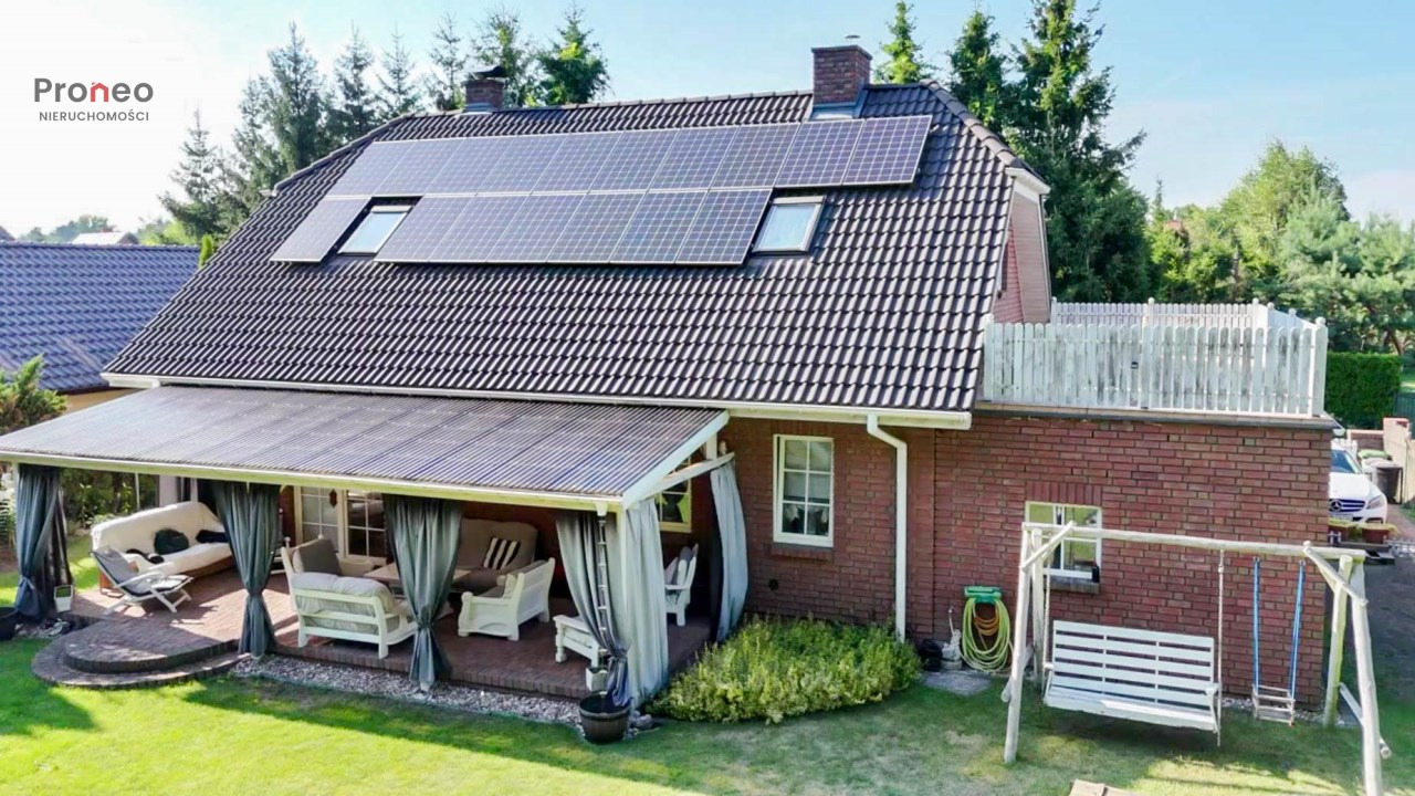 Dom z czerwonej cegły z panelami słonecznymi na dachu i zadaszonym patio z białymi meblami ogrodowymi. Huśtawka ogrodowa na pierwszym planie.