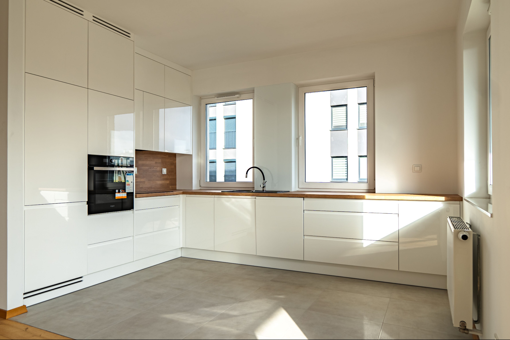 Jasna, nowoczesna kuchnia z białymi, lakierowanymi frontami, drewnianym blatem i wbudowanym piekarnikiem. Widok na okna z widokiem na budynek mieszkalny.