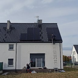 SABENERGY Sp. z O.O. - Montaż paneli fotowoltaicznych na dachu dwukondygnacyjnego domu z czarną dachówką; jeden instalator na drabinie, drugi na ziemi, widoczne są narzędzia i zabezpieczenia.