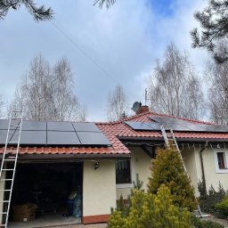 SABENERGY Sp. z O.O. - Dom jednorodzinny z zamontowanymi panelami fotowoltaicznymi na dachu z czerwonej dachówki, drabina oparta o dach, widoczne drzewa i krzewy w ogrodzie, pochmurne niebo.