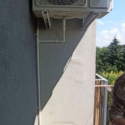 SABENERGY Sp. z O.O. - Zewnętrzna jednostka klimatyzacji Haier zamontowana na szarej ścianie budynku, widoczne białe rury odprowadzające skropliny i wiadro pod nimi. W tle zieleń drzew i fragment balkonu.