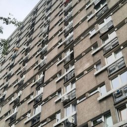 SABENERGY Sp. z O.O. - Wysoki blok mieszkalny z balkonami i oknami, widok z dołu, na jednym z balkonów widoczna osoba, na kilku balkonach zamontowane anteny satelitarne i klimatyzatory.