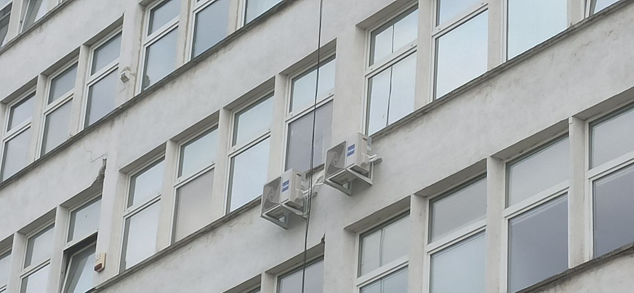 Dwa klimatyzatory zamontowane na elewacji budynku biurowego z widocznymi oknami i pęknięciem ściany w lewym górnym rogu. Widoczne przewody doprowadzające zasilanie.
