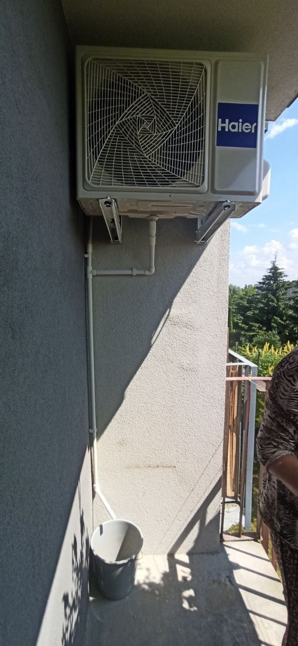Zewnętrzna jednostka klimatyzacji Haier zamontowana na szarej ścianie budynku, widoczne białe rury odprowadzające skropliny i wiadro pod nimi. W tle zieleń drzew i fragment balkonu.