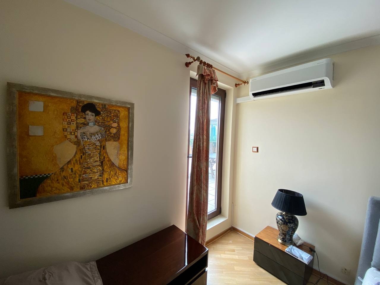 Biały klimatyzator zamontowany na ścianie nad oknem z dekoracyjnymi zasłonami, obok obraz Gustava Klimta i lampka nocna na szafce, w jasnym pokoju.