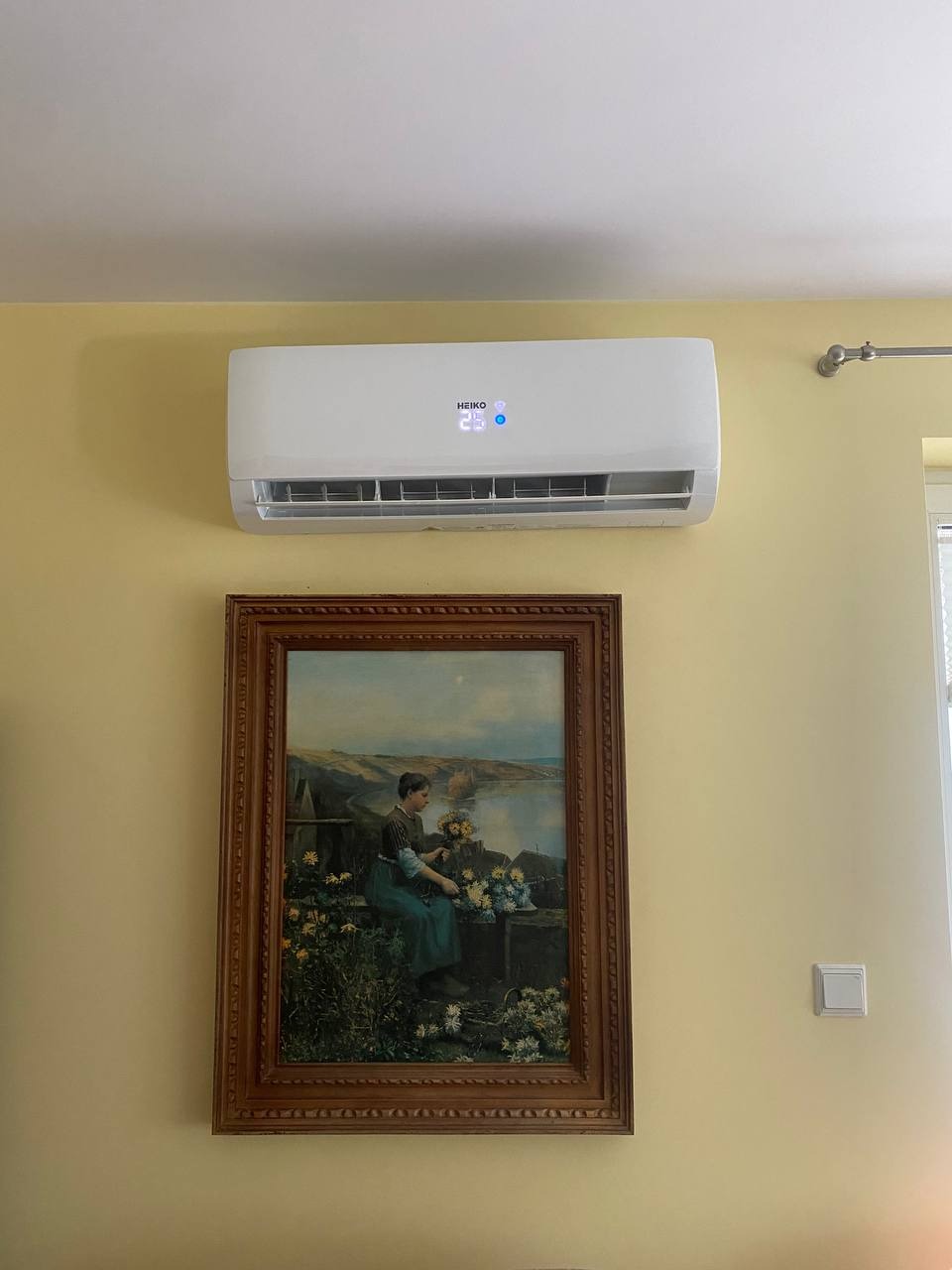 Biały klimatyzator z wyświetlaczem temperatury 25 stopni Celsjusza zamontowany nad obrazem w drewnianej ramie na żółtej ścianie.
