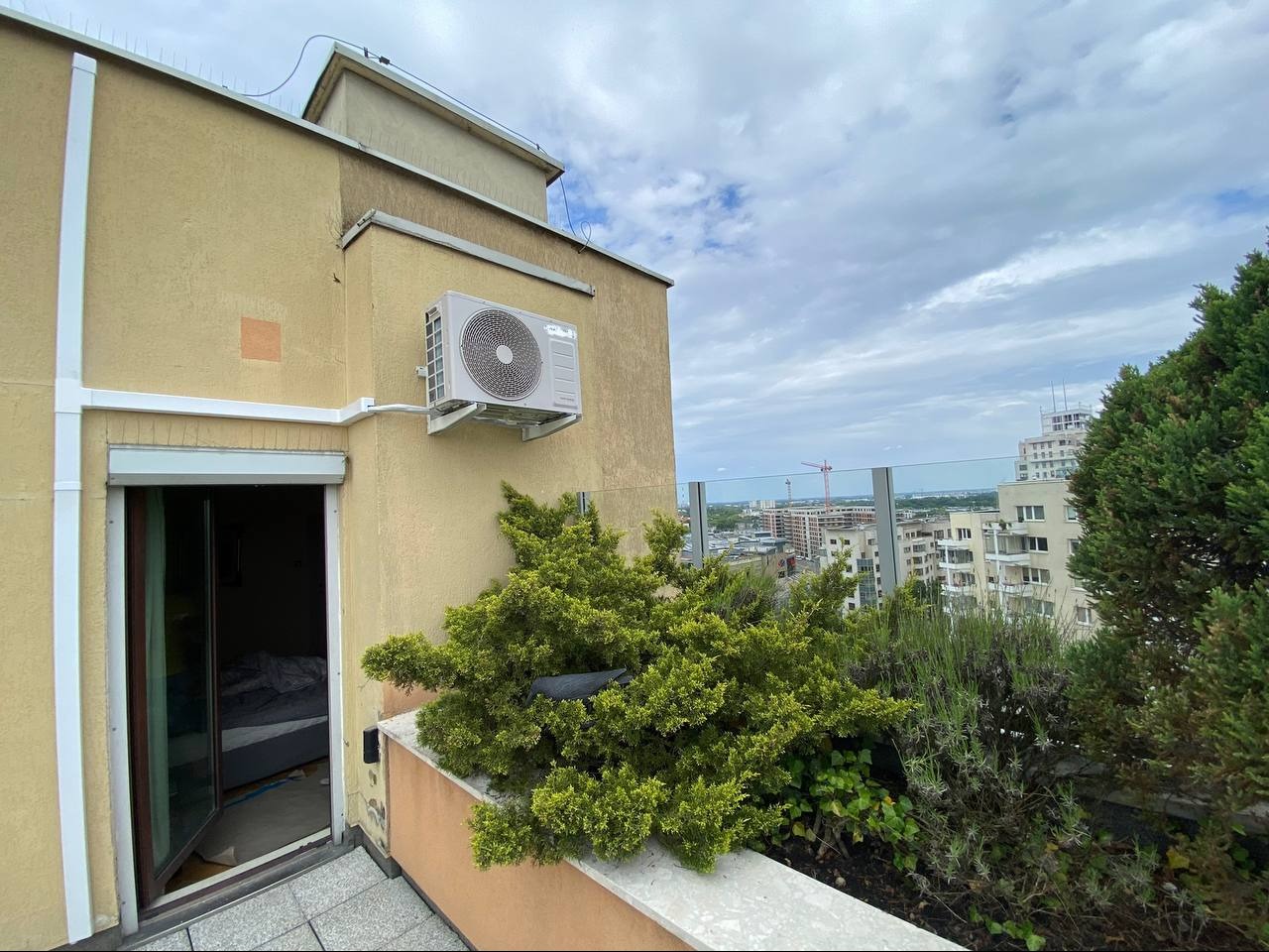 Balkon z widokiem na miasto, na ścianie budynku zamontowana jednostka zewnętrzna klimatyzacji, obok drzwi balkonowych rosną krzewy.