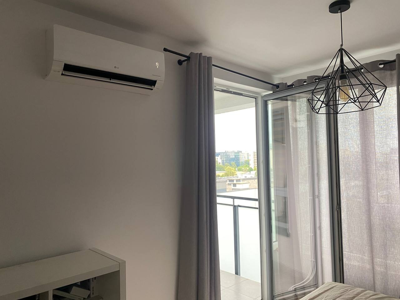 Nowoczesny klimatyzator LG zamontowany na ścianie nad białą półką, z widokiem na balkon i miasto w tle, zasłonięty częściowo przez szare zasłony i geometryczną lampę wiszącą.