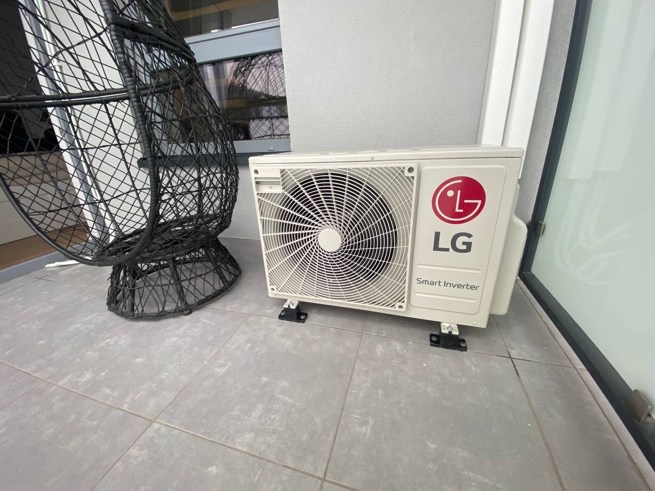 Zewnętrzna jednostka klimatyzacji LG Smart Inverter umieszczona na szarych płytkach tarasowych, obok czarnego, plecionego fotela wiszącego.