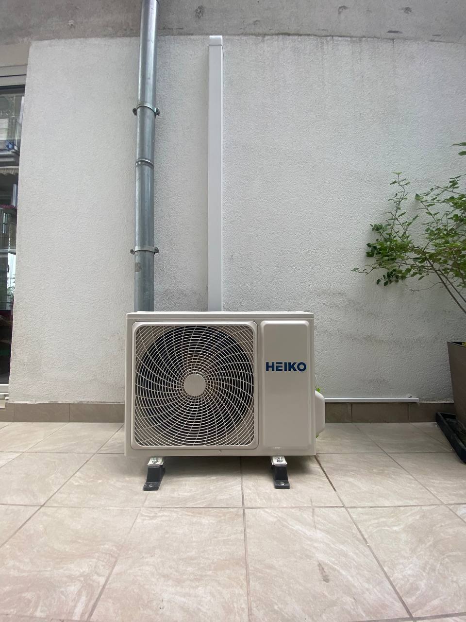 Biała jednostka zewnętrzna klimatyzatora marki Heiko, zamontowana na czarnych wspornikach na wyłożonym kafelkami balkonie, z widoczną rurą spustową i fragmentem rośliny w donicy w tle.