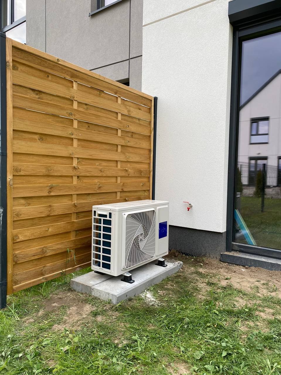 Biała jednostka zewnętrzna klimatyzacji/pompy ciepła, zamontowana na betonowej podstawie obok drewnianego ogrodzenia i ściany budynku z oknem.