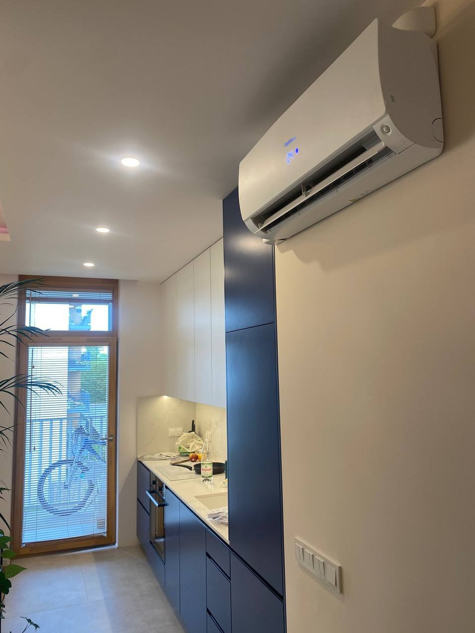 Biała klimatyzacja zamontowana na ścianie w nowoczesnej kuchni z granatowymi szafkami, widok z dołu, okno z widokiem na zewnątrz.