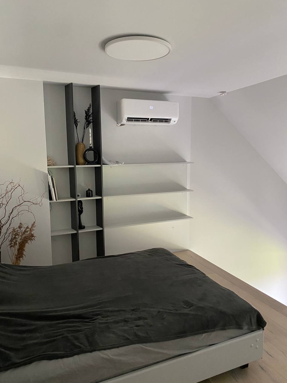 Klimatyzator ścienny zamontowany w sypialni nad półkami, widoczny z perspektywy łóżka z ciemną narzutą.