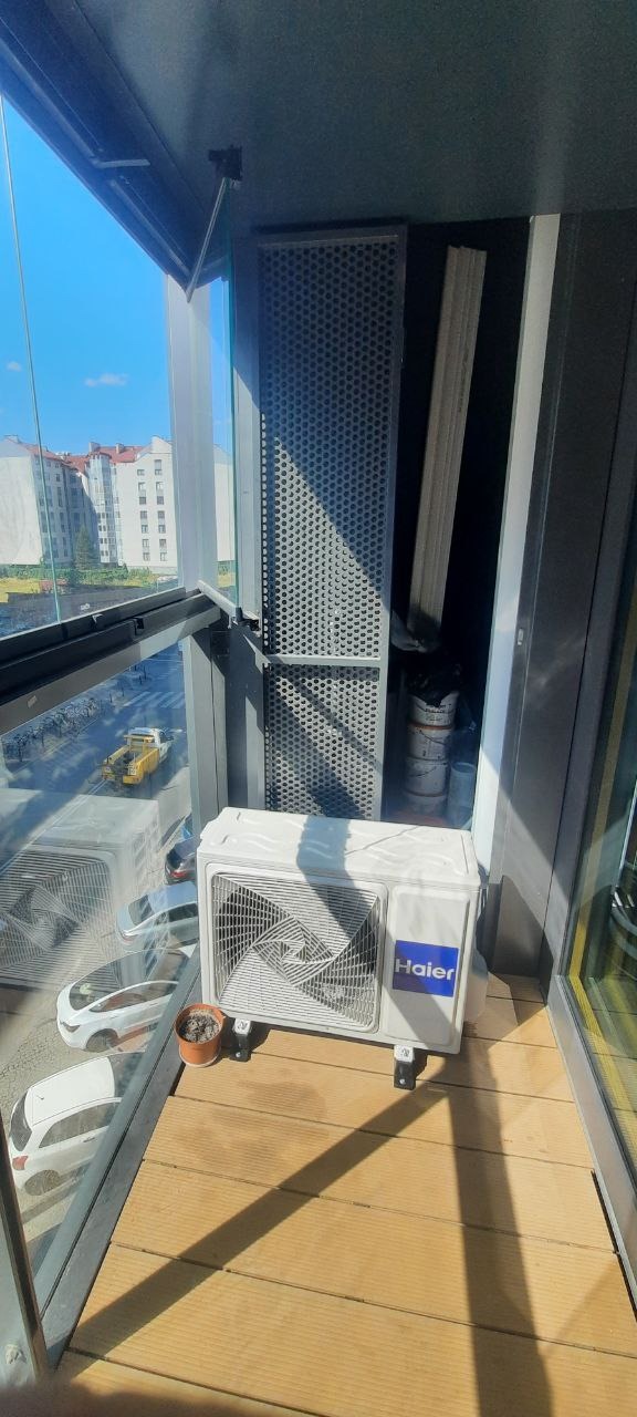 Kompaktowa jednostka zewnętrzna klimatyzacji marki Haier, ustawiona na drewnianej podłodze balkonu z widokiem na miejski krajobraz, obok mała doniczka z rośliną.