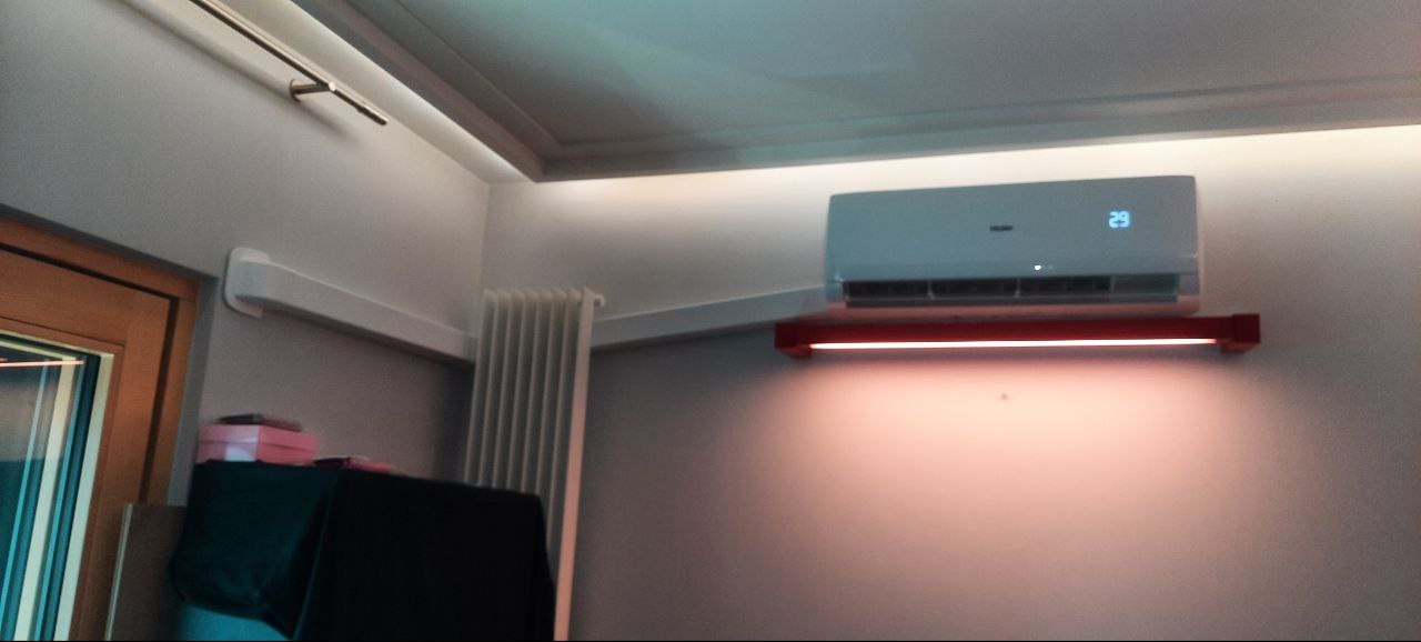 Klimatyzator Haier zamontowany na ścianie z czerwoną listwą oświetleniową, widoczny grzejnik i fragment drzwi z ościeżnicą.