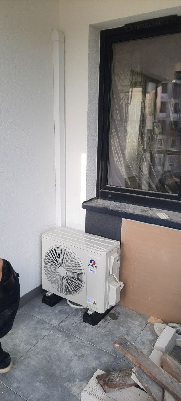 Biała jednostka zewnętrzna klimatyzacji marki Gree na balkonie, zamontowana na czarnych podstawkach, widoczna osoba w czarnych spodniach, okno zabezpieczone folią, materiały budowlane obok.