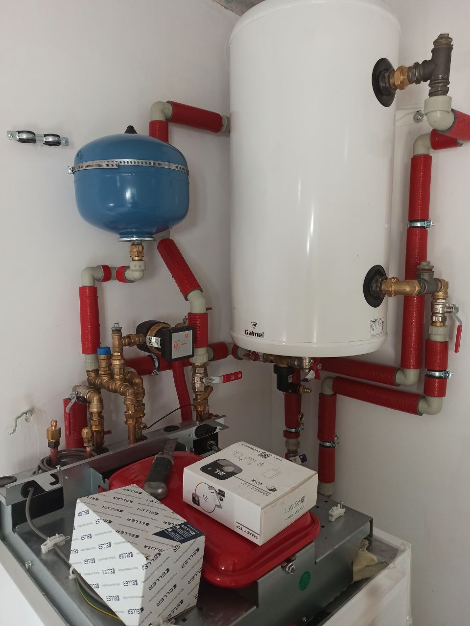 Złożony system instalacji hydraulicznej z białym zasobnikiem ciepłej wody, niebieskim naczyniem przeponowym, pompą obiegową i rurami w izolacji termicznej, dodatkowo widoczne pudełka z elementami...