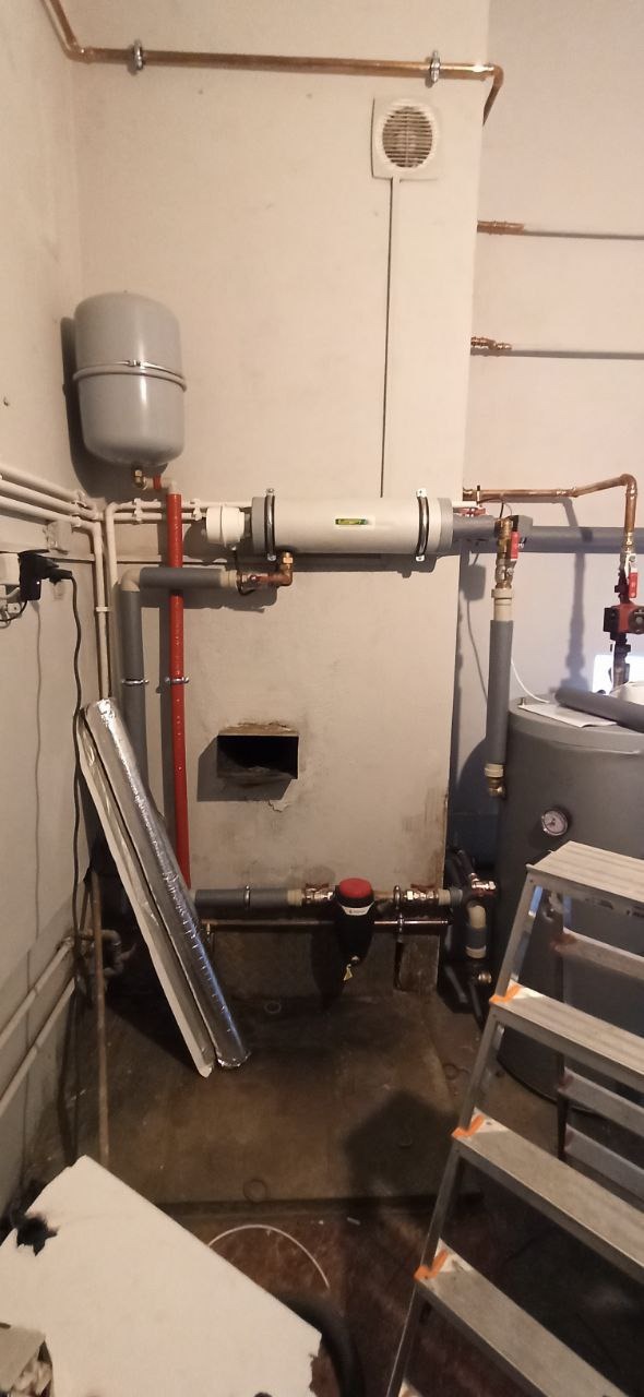 Kotłownia z zainstalowanym zbiornikiem wyrównawczym, filtrem, bojlerem i rurami miedzianymi, widoczny fragment drabiny i izolacji.