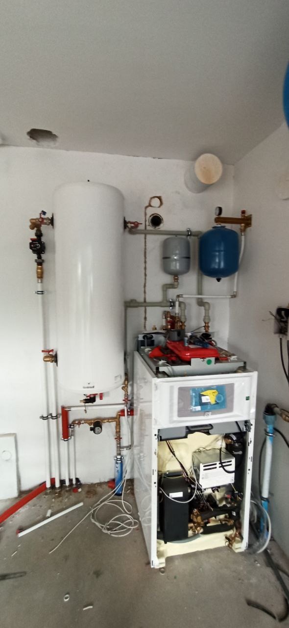 Biały bojler z podłączonymi rurami miedzianymi i zbiornikami wyrównawczymi w kotłowni w trakcie montażu, widoczne otwarte wnętrze pieca z przewodami, surowy stan wykończenia pomieszczenia.