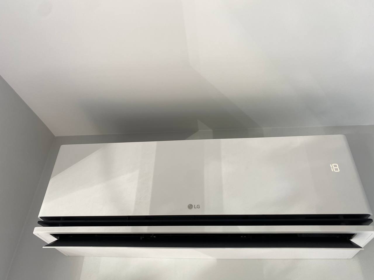 Biały klimatyzator LG zamontowany na ścianie, wyświetlający temperaturę 18 stopni, widoczny z dołu pod kątem.