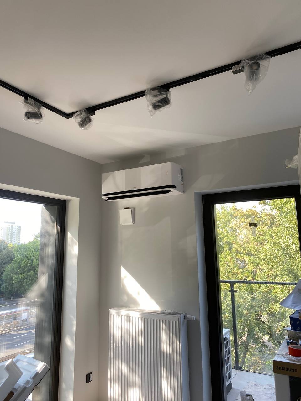 Wnętrze pokoju z zamontowanym klimatyzatorem LG na ścianie, grzejnikiem panelowym pod oknem, reflektorami sufitowymi w folii ochronnej na czarnej szynie oraz kratką wentylacyjną na ścianie.