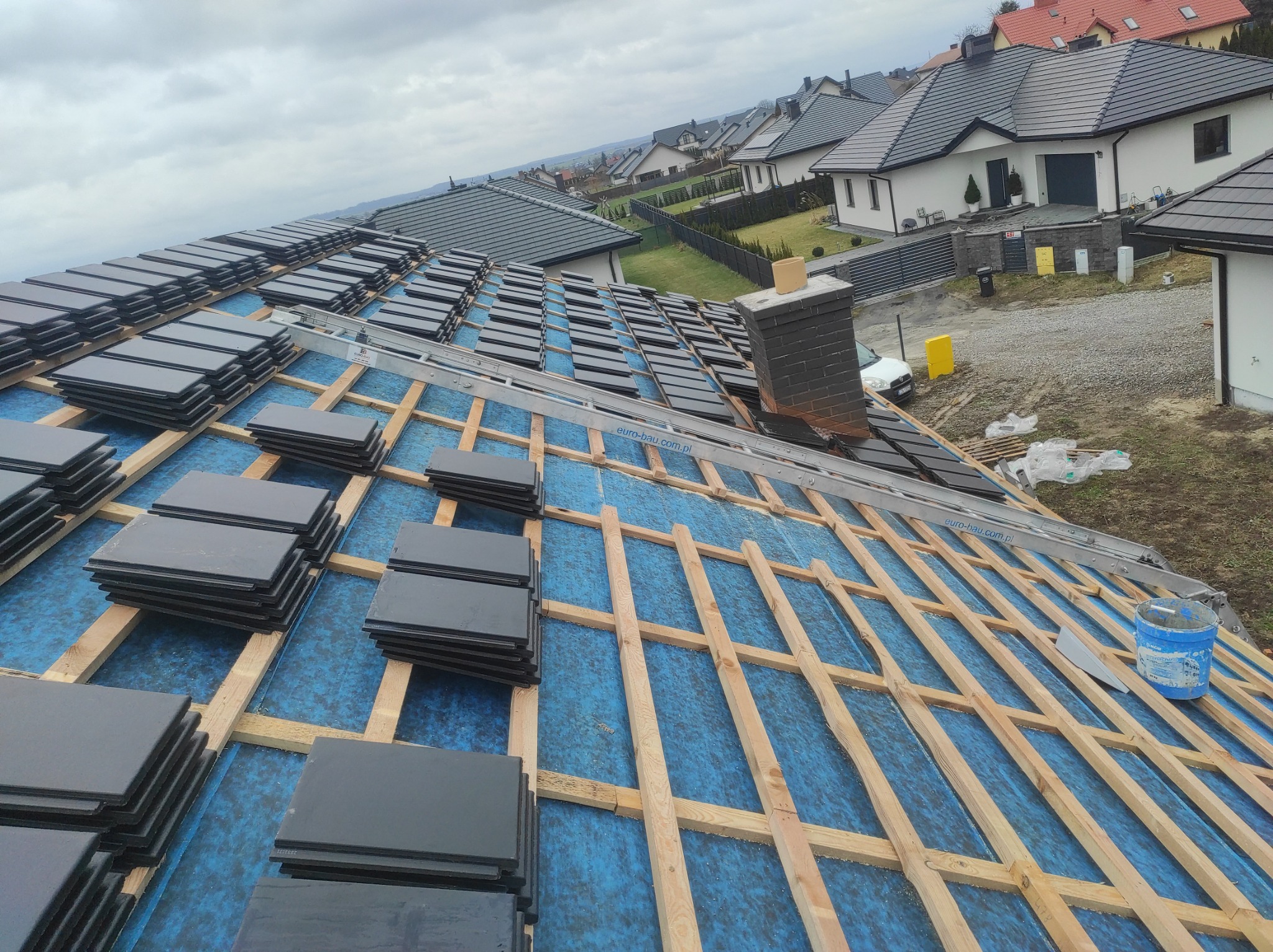 Widok z góry na rozłożone dachówki na konstrukcji drewnianej pokrytej niebieską membraną, przygotowane do ułożenia na dachu domu jednorodzinnego na tle zabudowy mieszkalnej w pochmurny dzień.