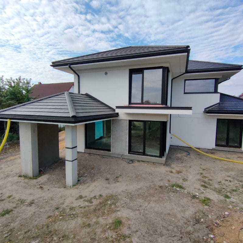 Nowoczesny dom jednorodzinny z szarą dachówką karpiówką i czarnymi oknami, widoczne rynny w kolorze dachu, teren wokół domu jeszcze niezagospodarowany, na pierwszym planie zadaszenie nad wejściem...