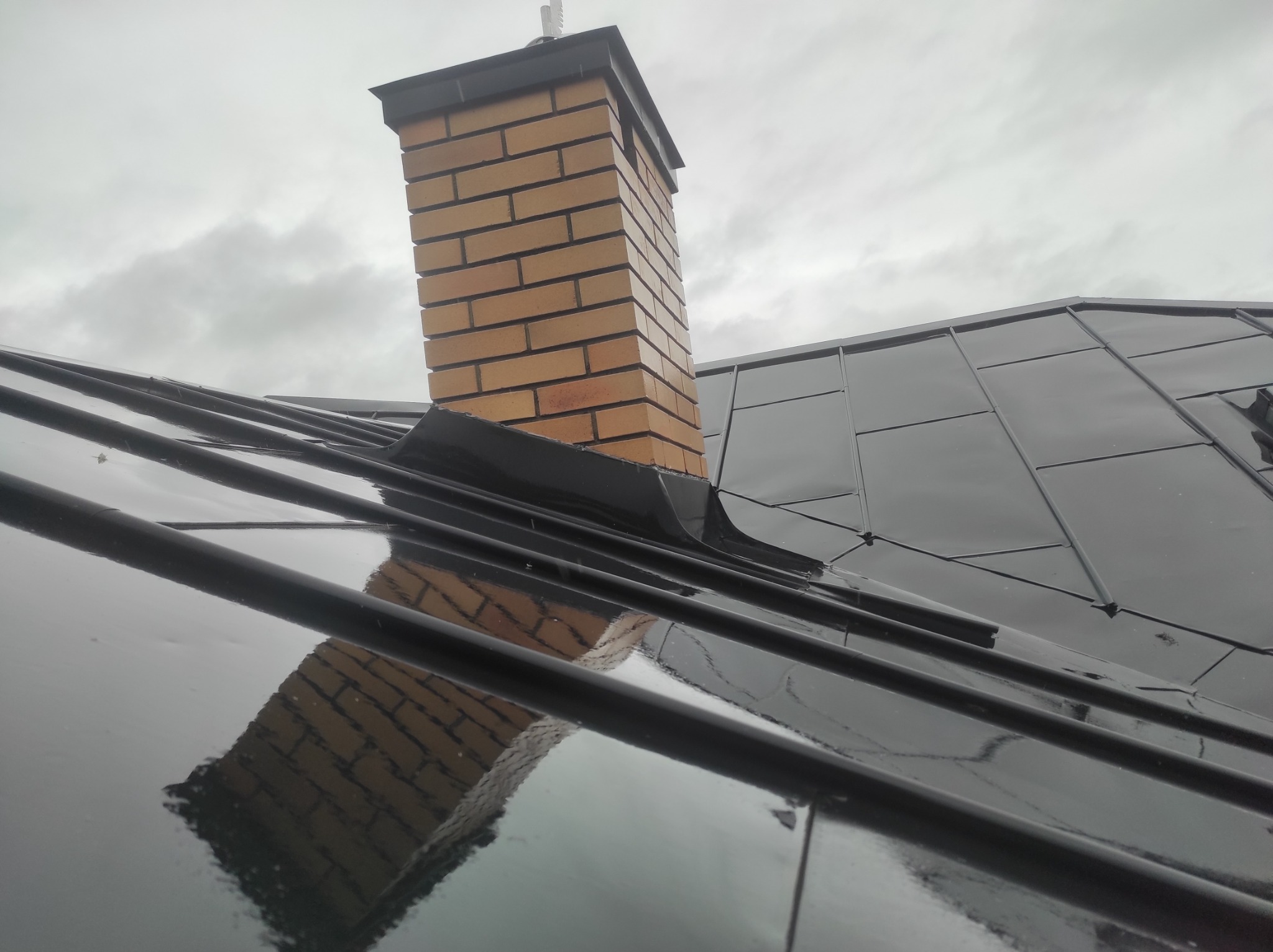 Błyszczący, czarny dach z metalowymi panelami i ceglanym kominem w pochmurny dzień. Widoczne refleksy komina na powierzchni dachu.