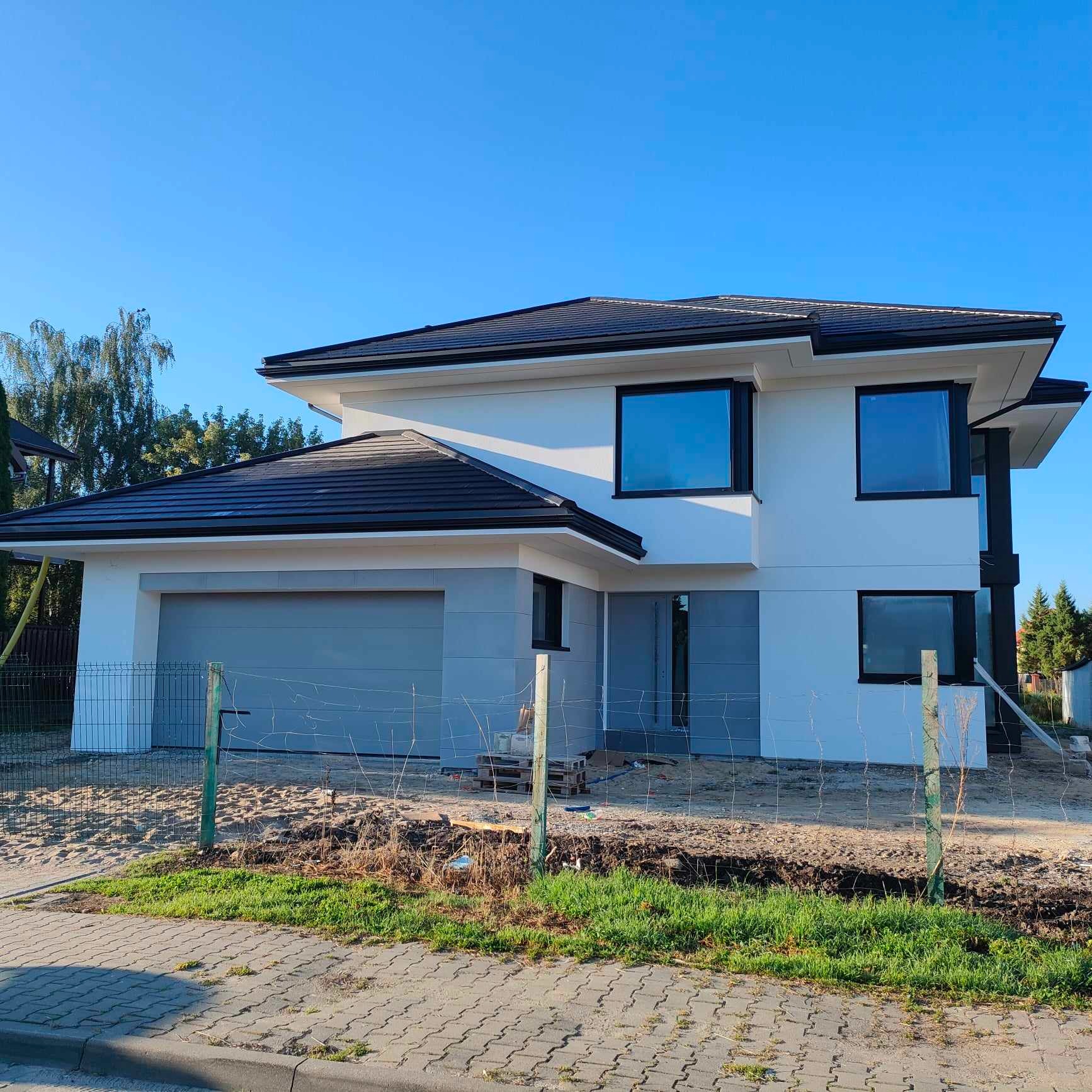 Nowoczesny dom jednorodzinny z ciemną dachówką, szarą bramą garażową i białymi ścianami, otoczony tymczasowym ogrodzeniem z siatki.