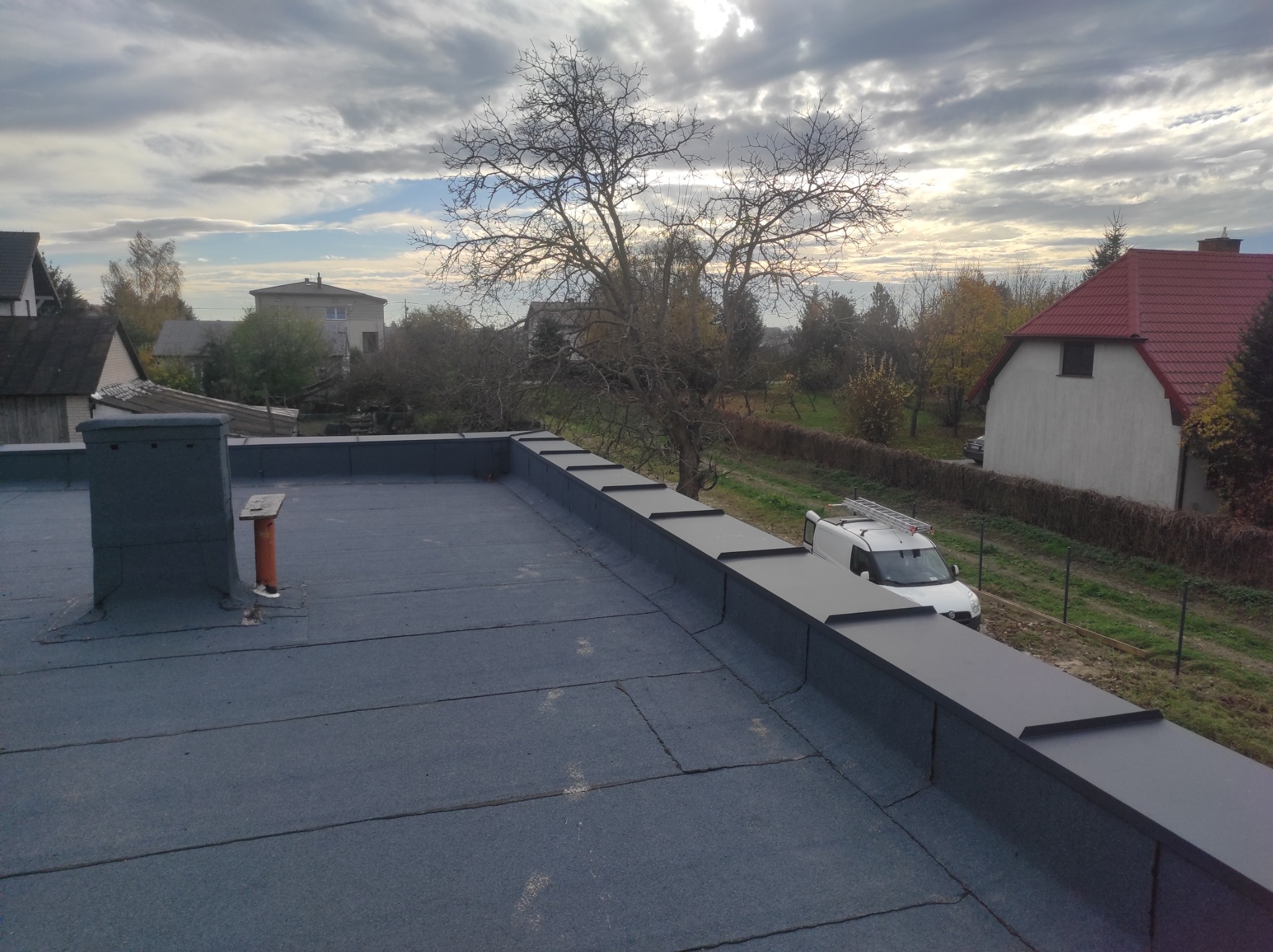 Widok z góry na płaski dach pokryty papą, z widocznymi obróbkami blacharskimi na krawędziach i kominem, w tle zabudowa jednorodzinna i samochód dostawczy z drabiną na dachu.