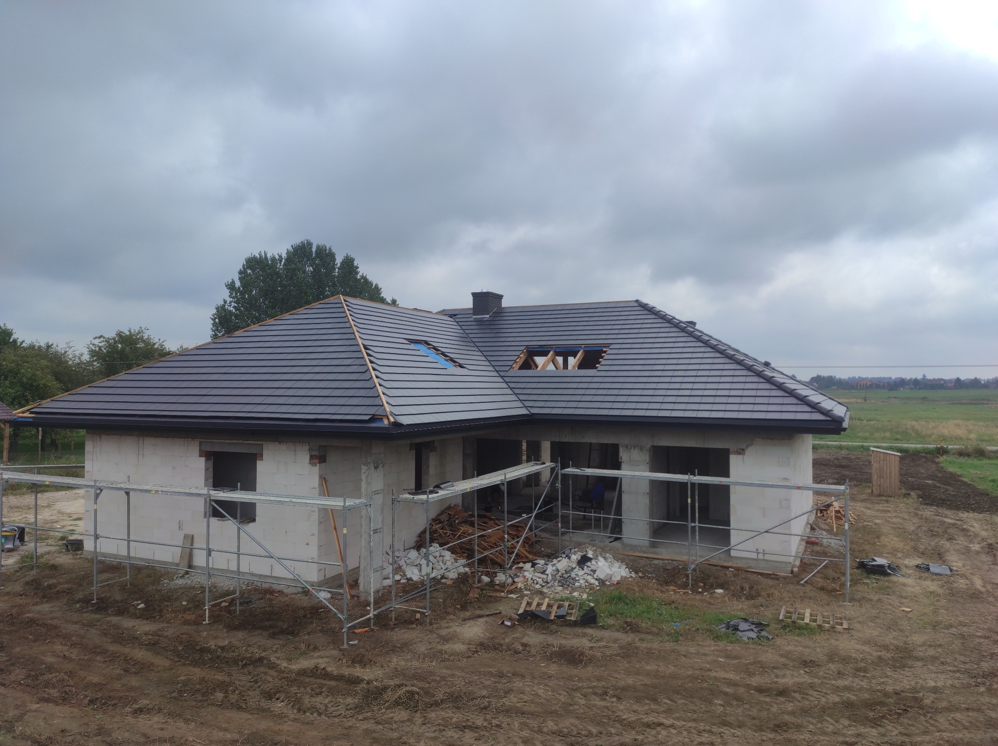 Dom jednorodzinny w trakcie budowy, z częściowo położonym, grafitowym dachem z dachówki ceramicznej, widoczne rusztowania i materiały budowlane na posesji, pochmurne niebo.