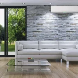 Biała klimatyzacja ścienna w nowoczesnym salonie z kamienną ścianą i jasną sofą narożną, widok na zieleń za oknem.