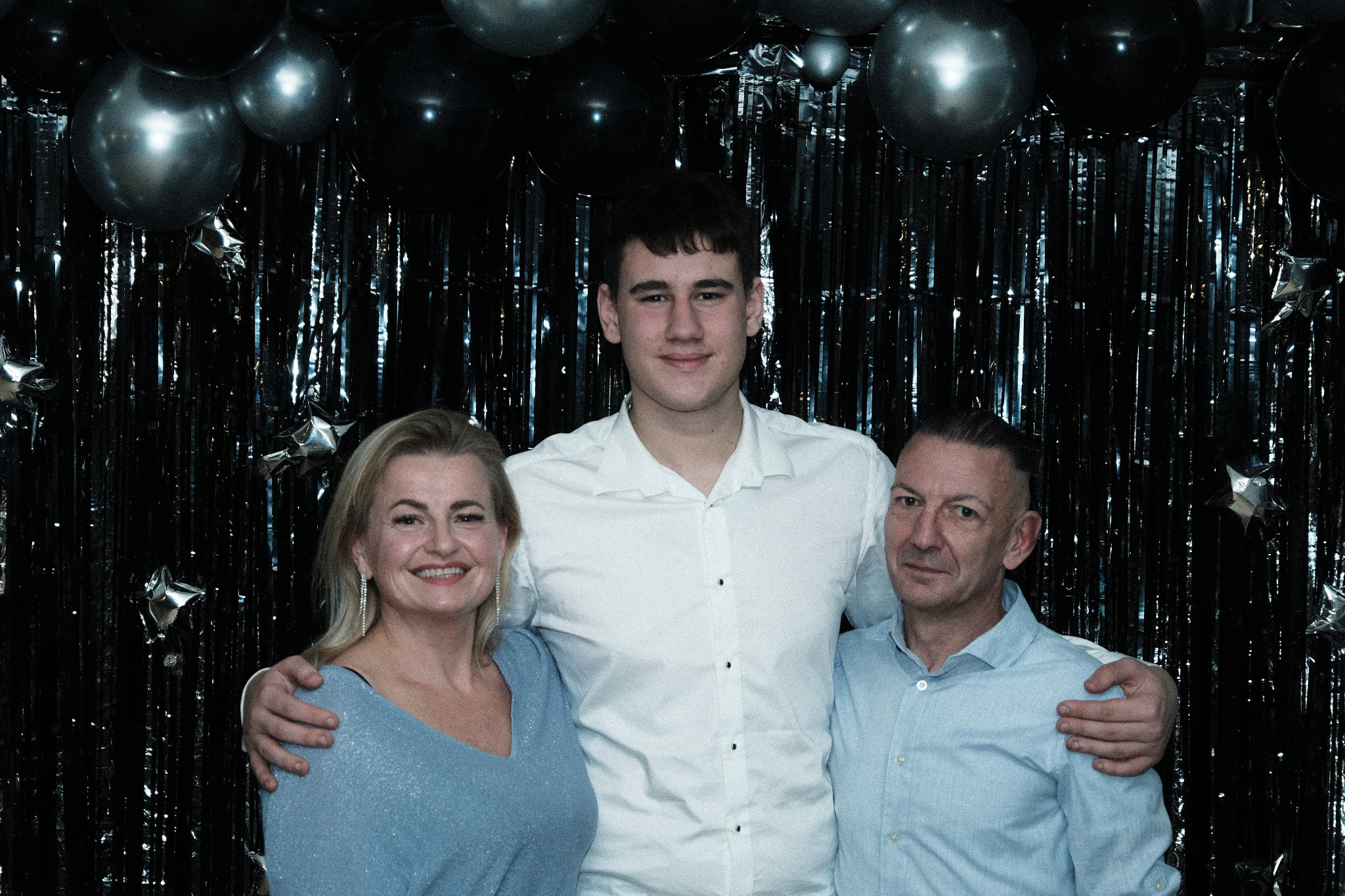 Portret rodzinny w Szczecinie: uśmiechnięta kobieta w niebieskiej sukience, wysoki chłopak w białej koszuli i mężczyzna w błękitnej koszuli, pozują na tle błyszczącej, czarnej kurtyny i srebrnych...