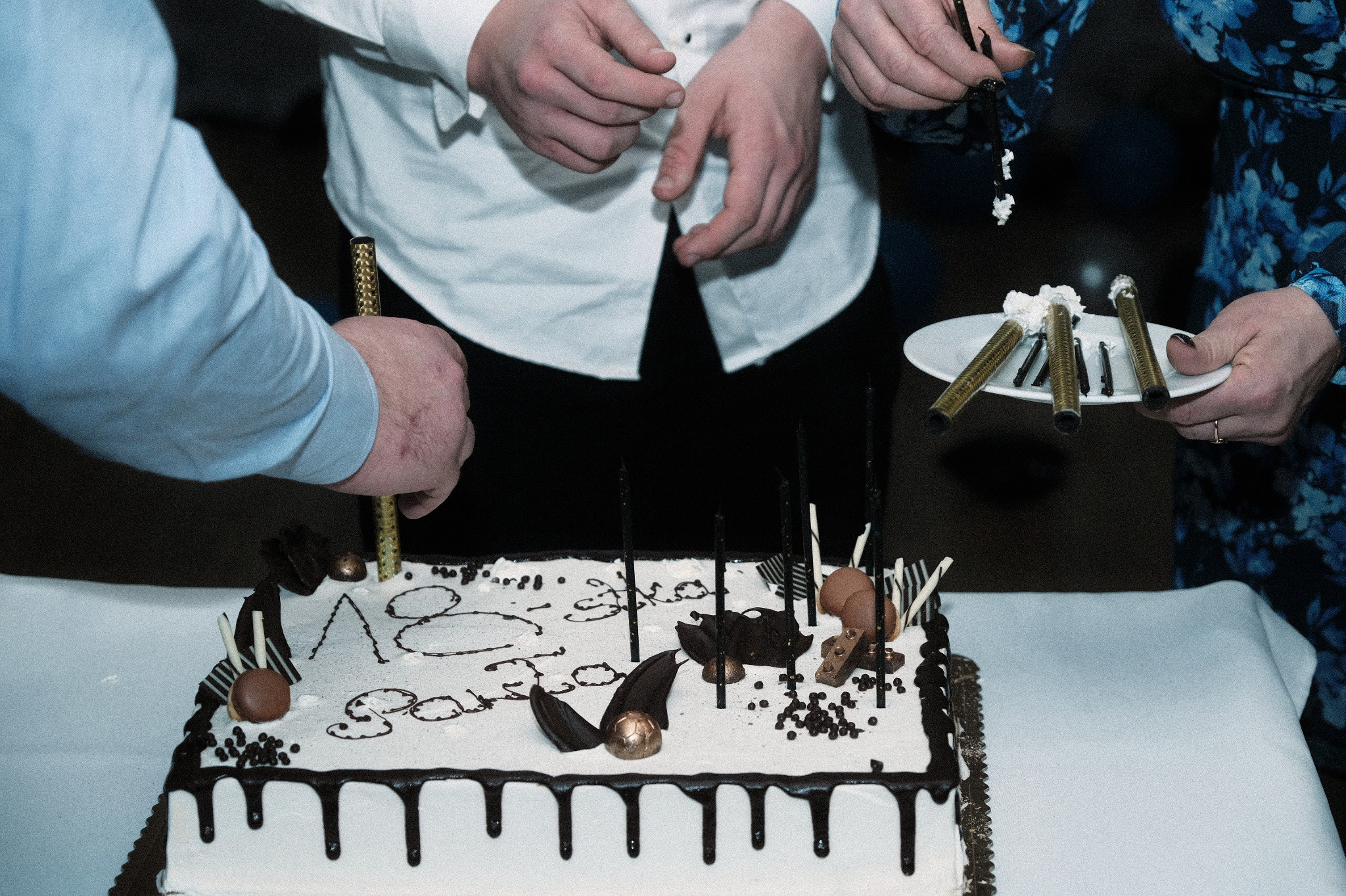 Udekorowany tort urodzinowy z czekoladowymi ozdobami i świeczkami, przygotowywany przez trzy osoby w białych i niebieskich koszulach, jedna z nich umieszcza złotą świeczkę na torcie, inna trzyma...