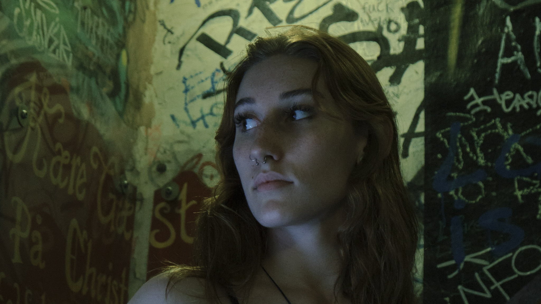 Portret młodej kobiety z piercingiem w nosie, spoglądającej w lewo, na tle ściany pokrytej graffiti w odcieniach zieleni, brązu i czerni.