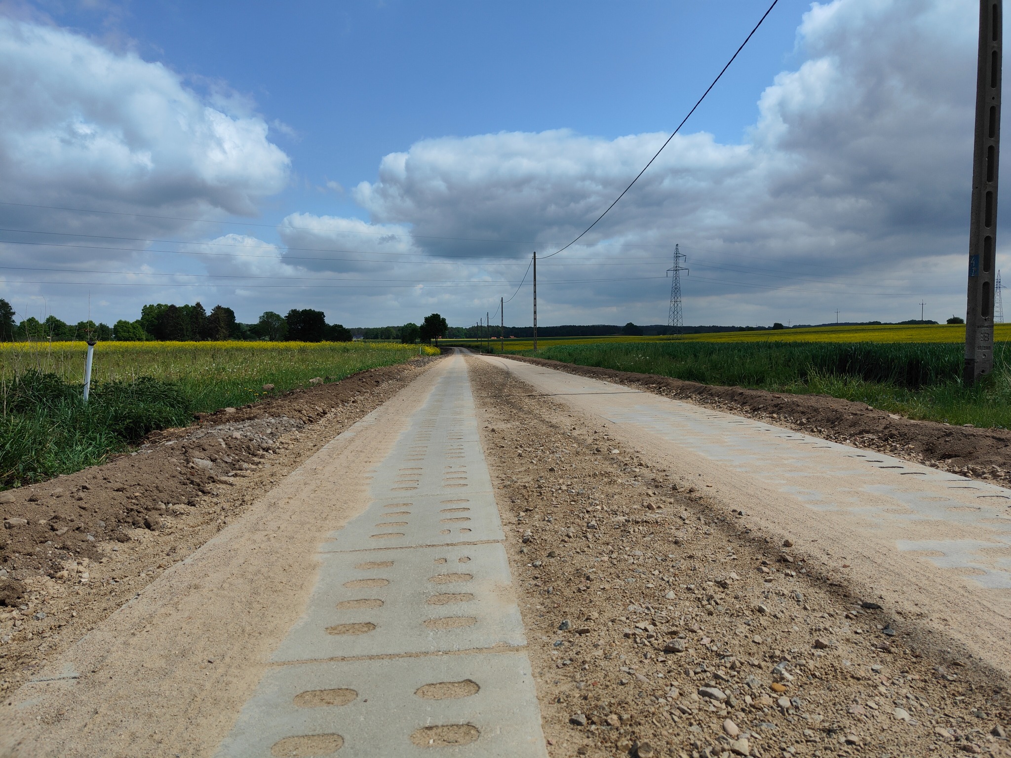Długa droga gruntowa wzmocniona prefabrykowanymi elementami betonowymi, prowadząca przez pola, z widocznymi słupami energetycznymi i chmurami na niebie.