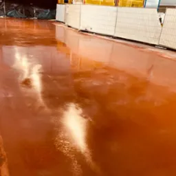 Świeżo wylana, błyszcząca, pomarańczowa posadzka przemysłowa z widocznymi refleksami światła, zabezpieczona tymczasowymi panelami ogrodzeniowymi w tle.