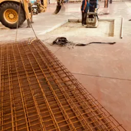 Wnętrze hali w Tychach: widok na przygotowane zbrojenie, pracownik zagęszcza podłoże ubijarką, w tle koparko-ładowarka.