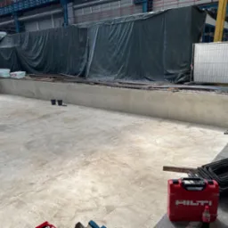 Wnętrze hali produkcyjnej z betonowym fundamentem w trakcie budowy, z widocznym zbrojeniem, narzędziami Hilti i zabezpieczeniem folią. Część podłoża betonowa, gotowa do dalszych prac.