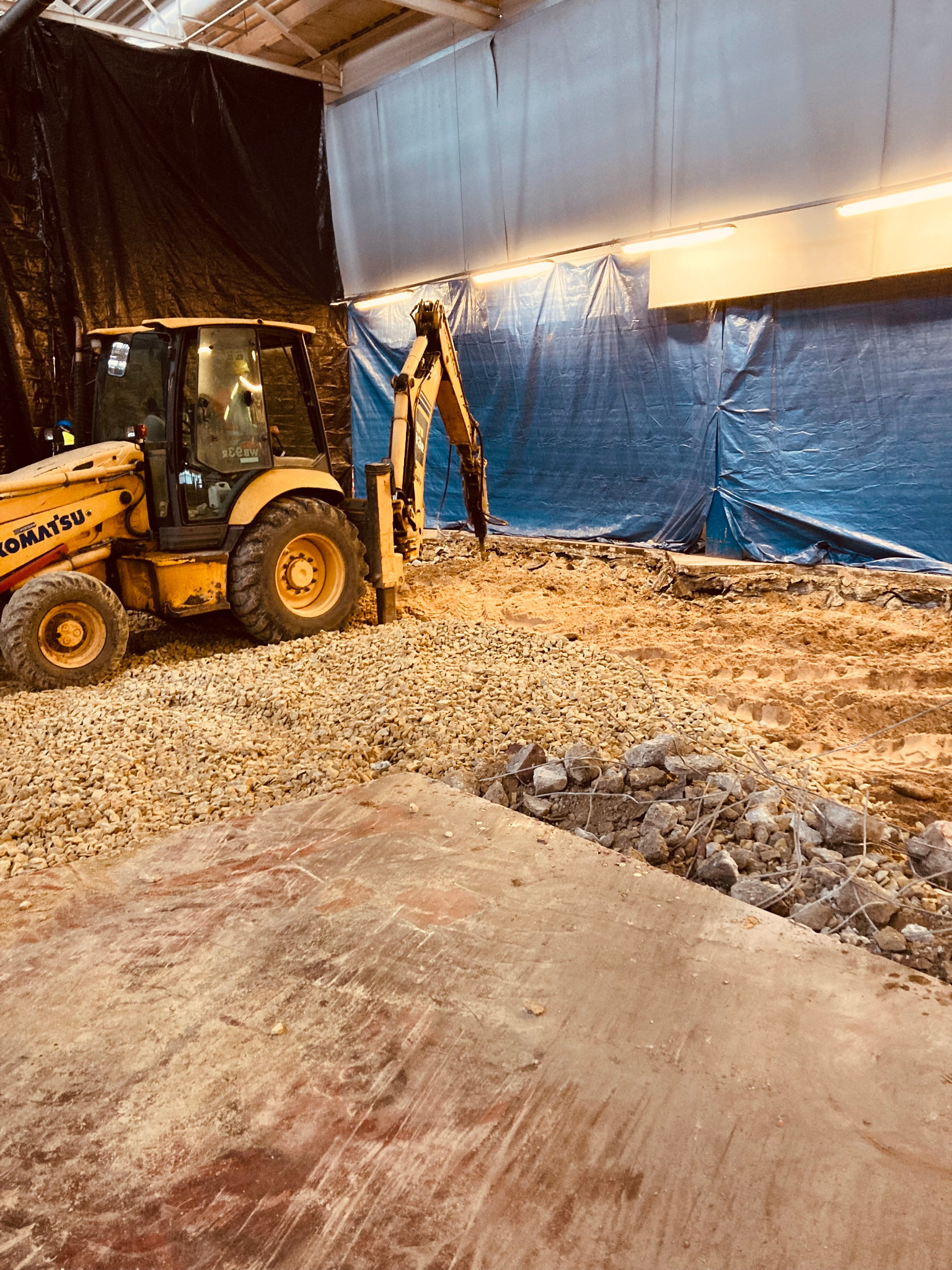 Żółta koparko-ładowarka Komatsu na placu budowy, widoczne prace ziemne i przygotowanie podłoża pod instalacje wewnątrz hali, z gruzem i kamieniami.