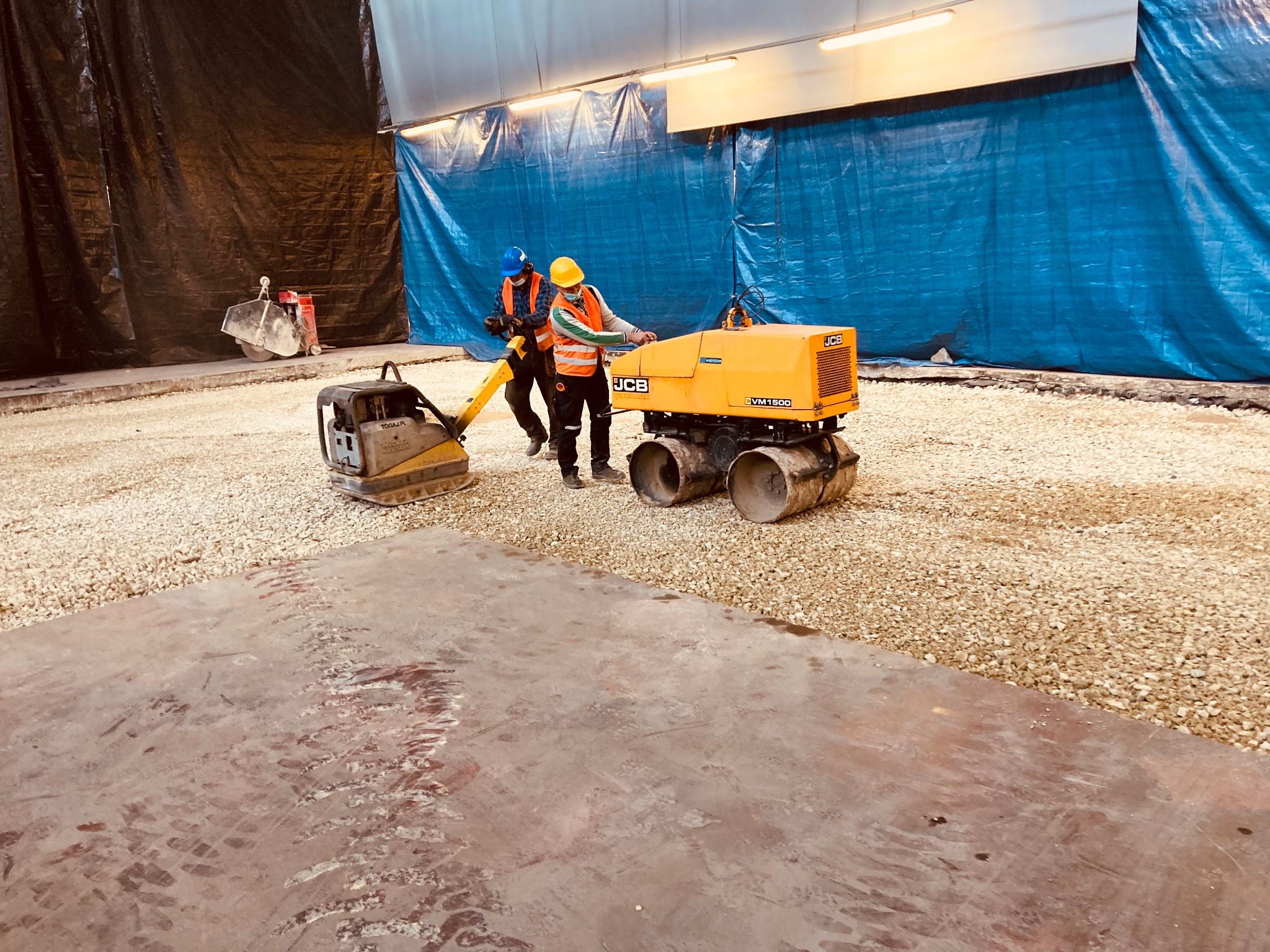 Dwaj pracownicy w kaskach i kamizelkach odblaskowych, używają zagęszczarki stopowej i walca wibracyjnego JCB VM150D do utwardzania podłoża z kruszywa pod posadzkę przemysłową w hali.