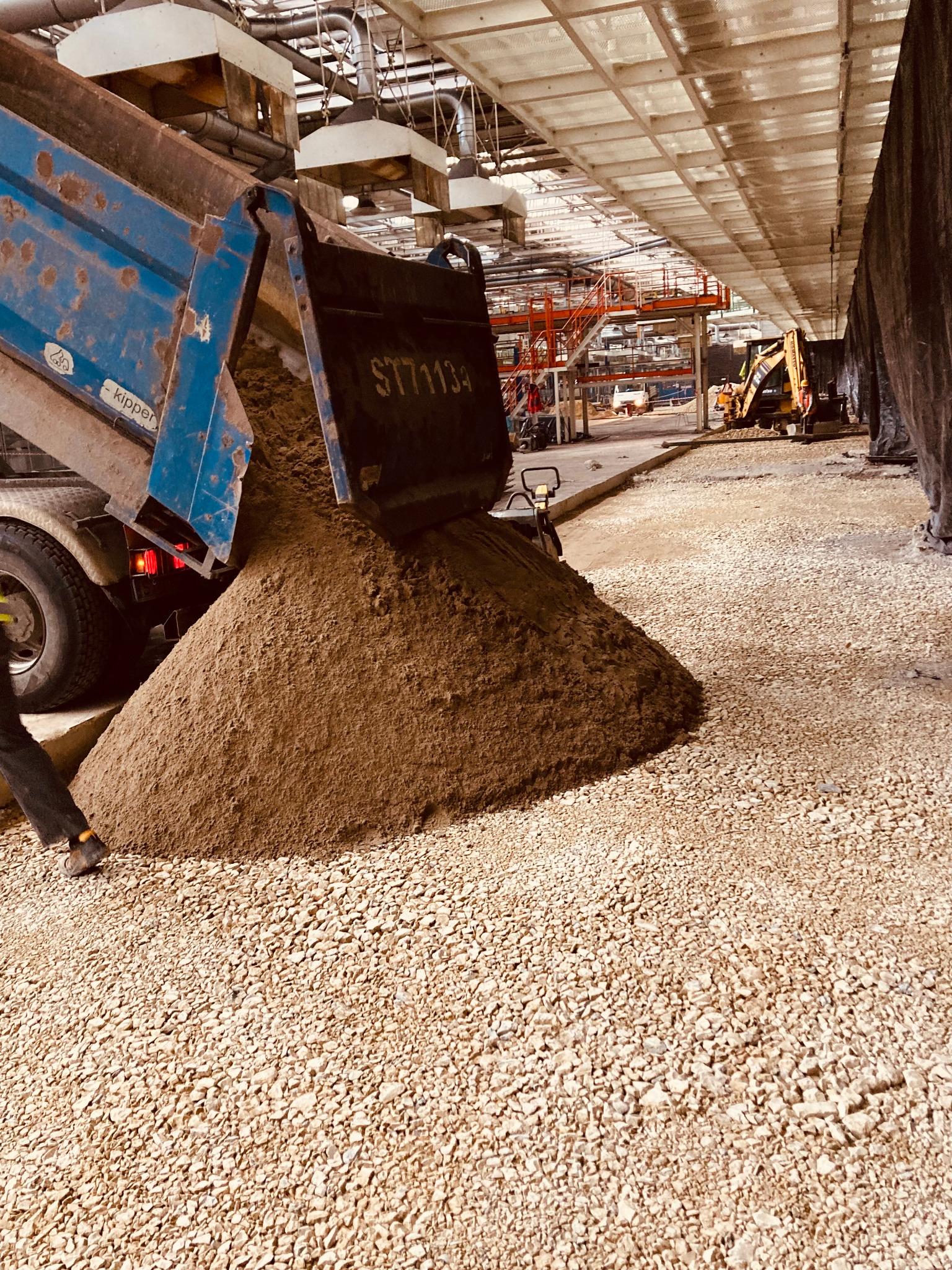 Wyładunek piasku z wywrotki na utwardzoną drogę z kruszywa, w tle widoczny sprzęt budowlany i konstrukcja hali.
