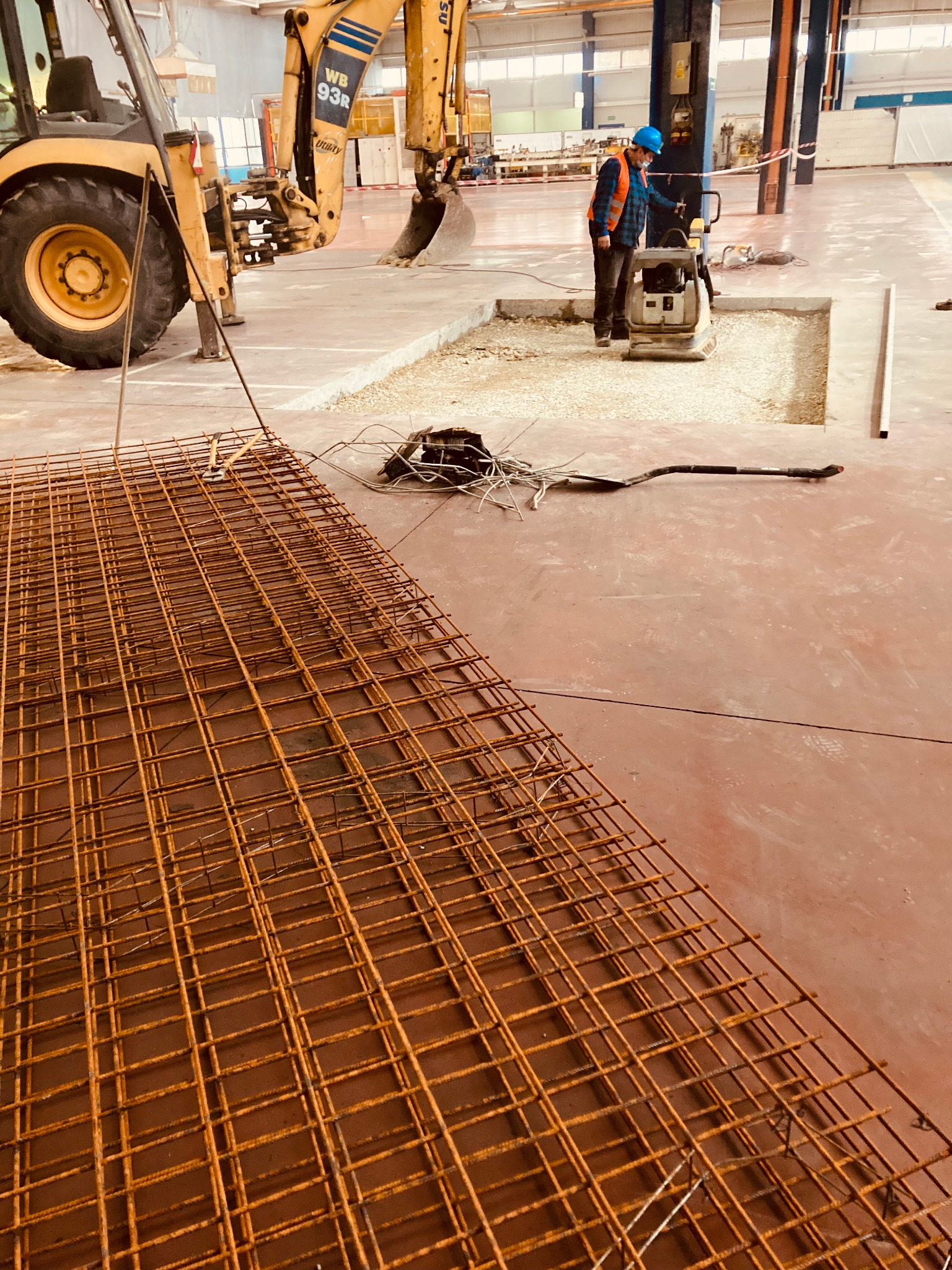 Wnętrze hali w Tychach: widok na przygotowane zbrojenie, pracownik zagęszcza podłoże ubijarką, w tle koparko-ładowarka.
