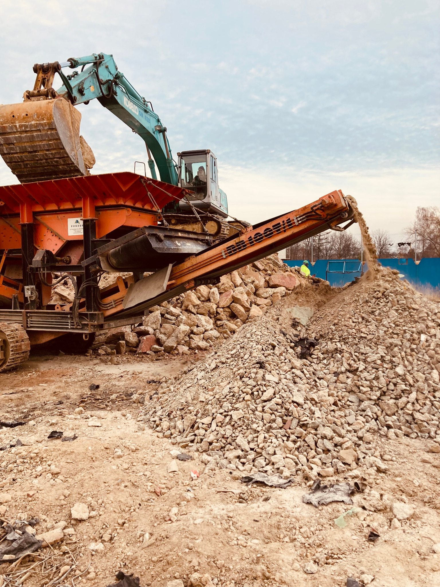 Ciężka maszyna budowlana przesiewa gruz na placu budowy, z operatorem w kabinie i pracownikiem w odblaskowej kamizelce w tle.