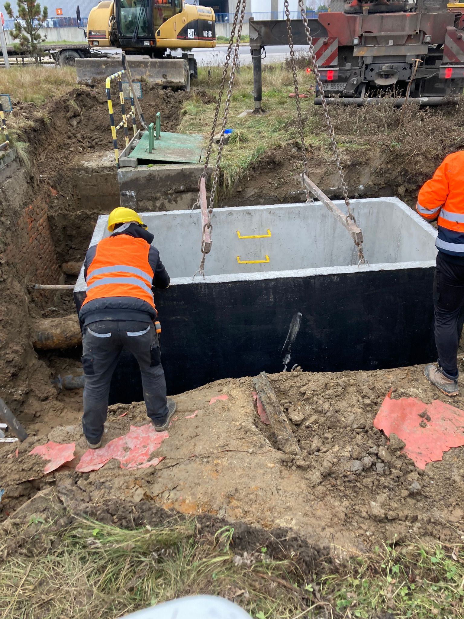 Osadzanie prefabrykowanego, betonowego zbiornika w wykopie. Widoczne stalowe liny dźwigu, robotnik w żółtym kasku i pomarańczowej kamizelce oraz wykop z widoczną instalacją rurową.