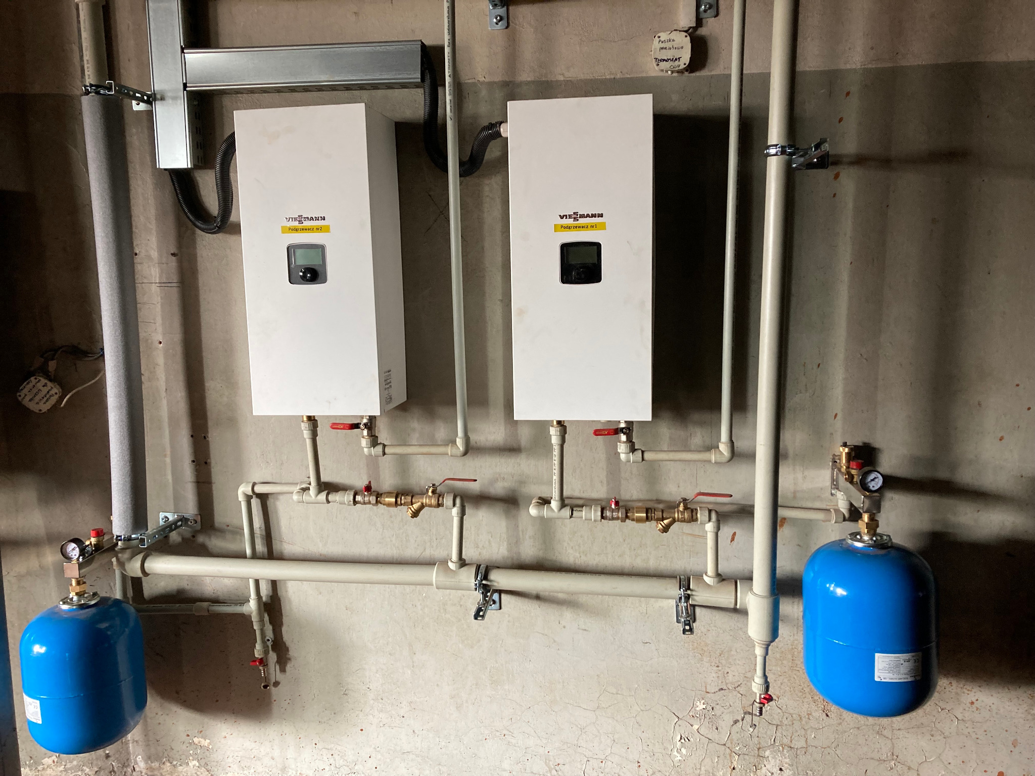 Dwa białe kotły grzewcze Viessmann z podłączonymi rurami i niebieskimi zbiornikami ciśnieniowymi zamontowane na szarej, popękanej ścianie w pomieszczeniu technicznym.