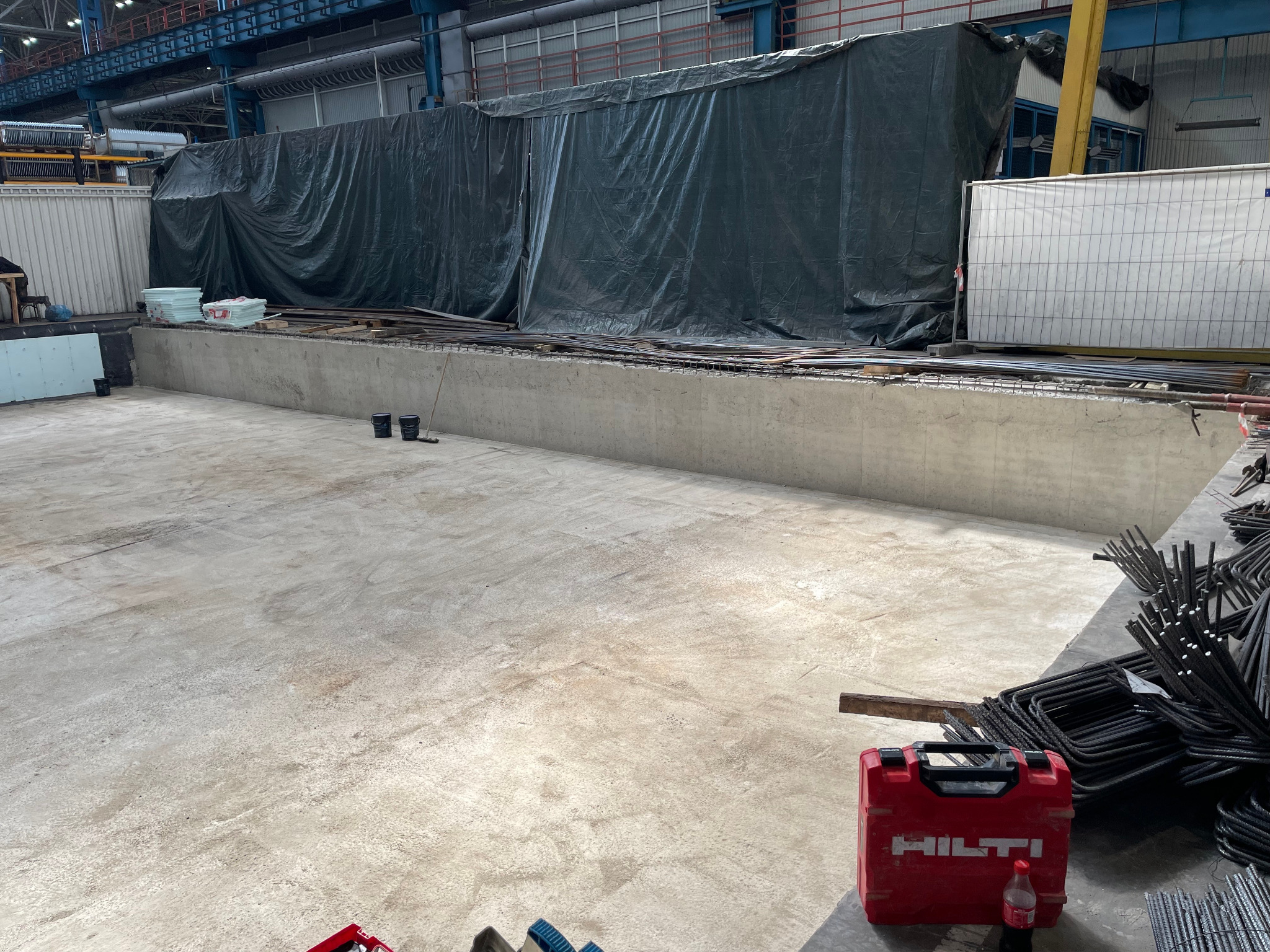 Wnętrze hali produkcyjnej z betonowym fundamentem w trakcie budowy, z widocznym zbrojeniem, narzędziami Hilti i zabezpieczeniem folią. Część podłoża betonowa, gotowa do dalszych prac.