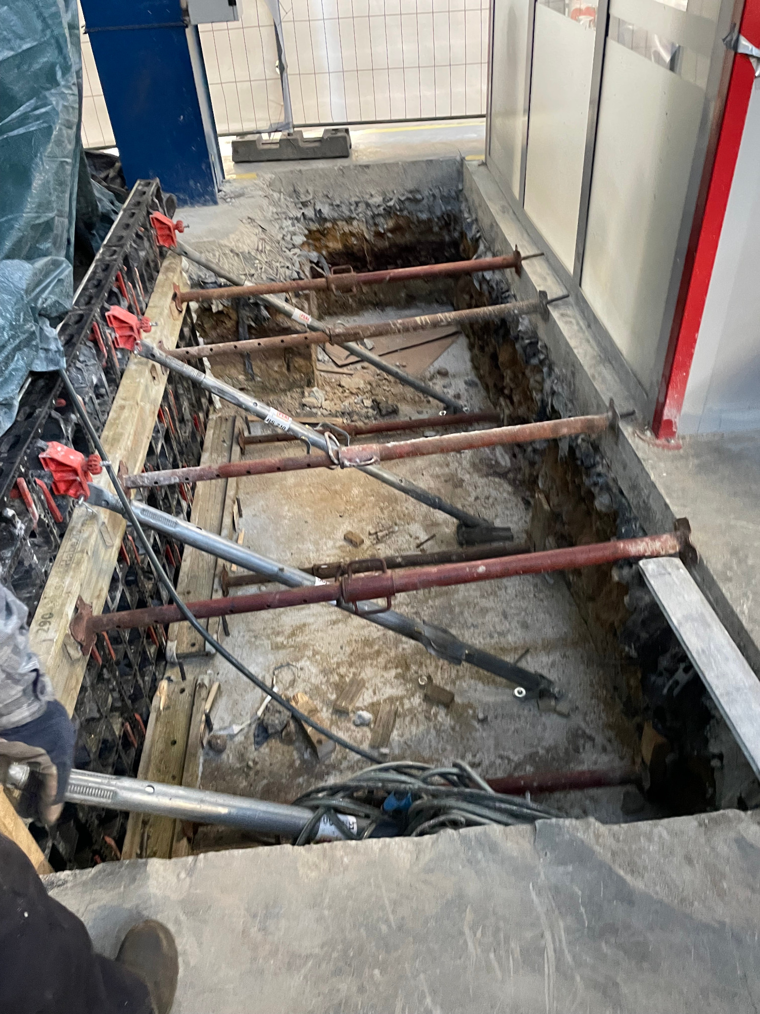 Wykop zabezpieczony szalunkiem i stemplami stalowymi, widoczne kable elektryczne i fragment zbrojenia.