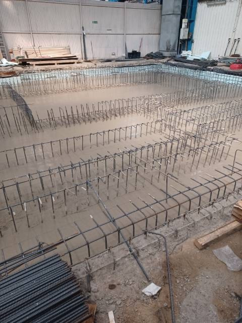 Zbrojenie fundamentów płyty betonowej z widocznymi prętami stalowymi i deskowaniem, przygotowane do zalania betonem, na tle magazynu z blachy falistej.