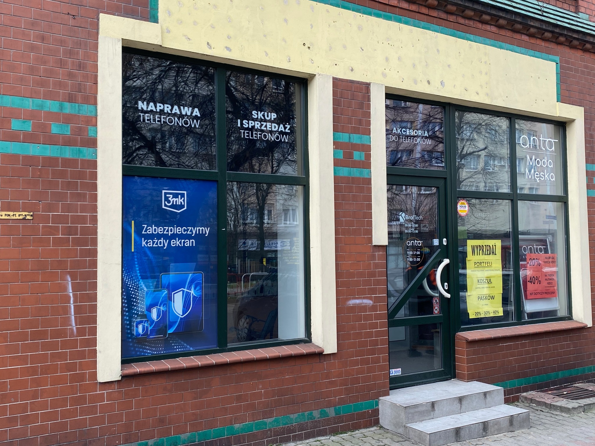 Fasada budynku z witrynami reklamującymi usługi: naprawa i skup telefonów, akcesoria do telefonów oraz moda męska. Na szybach naklejki z logo 3mk i informacją o zabezpieczaniu każdego ekranu.